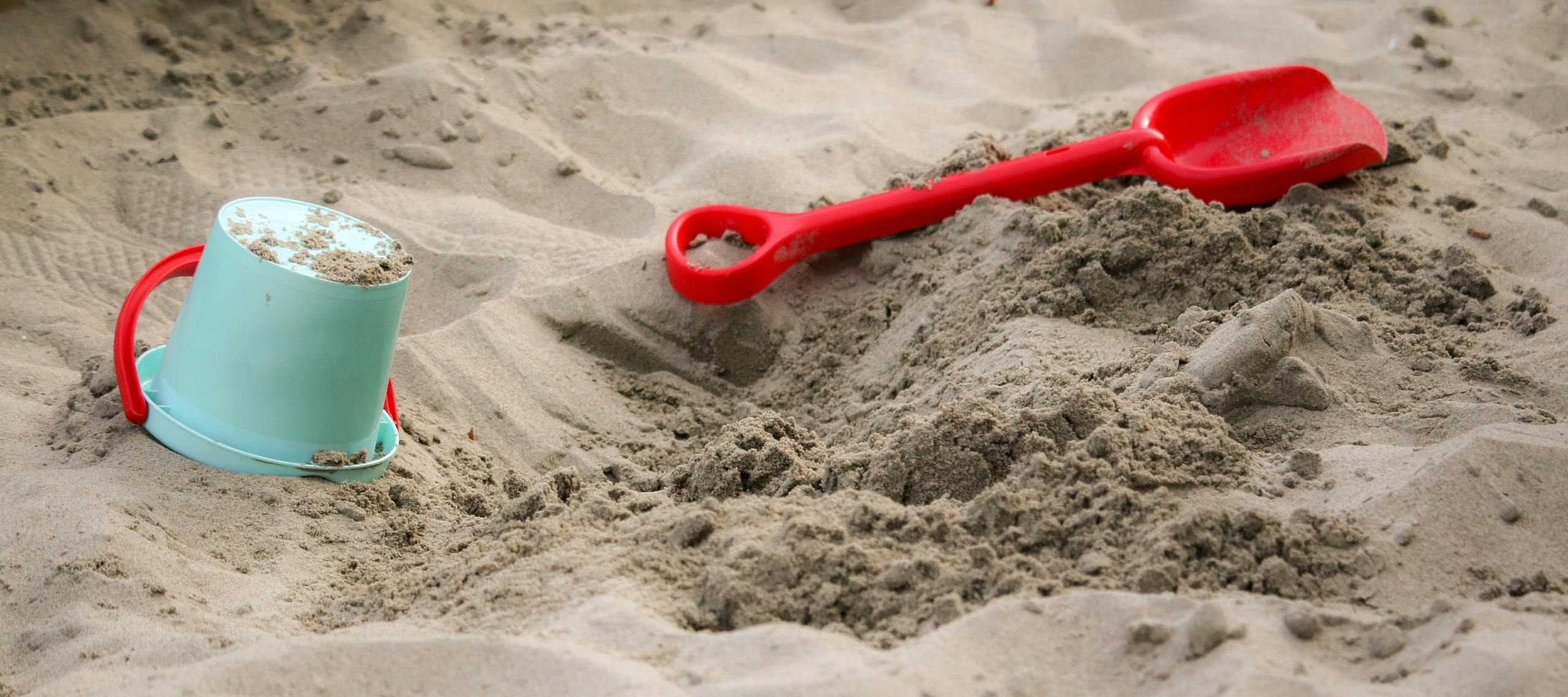 Red shovel and blue bucket in sand on a sunny beach day.